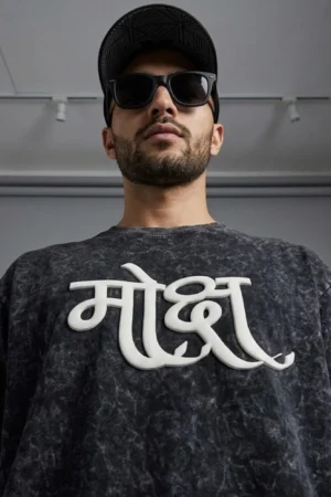 Oversized black acid wash t-shirt with 3D puff print Hindi "Moksha" (मोक्ष) calligraphy graphic in white, hanging on a wooden hanger against a beige wall background