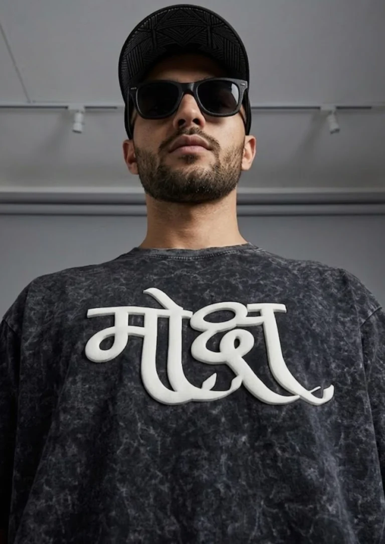 Oversized black acid wash t-shirt with 3D puff print Hindi "Moksha" (मोक्ष) calligraphy graphic in white, hanging on a wooden hanger against a beige wall background