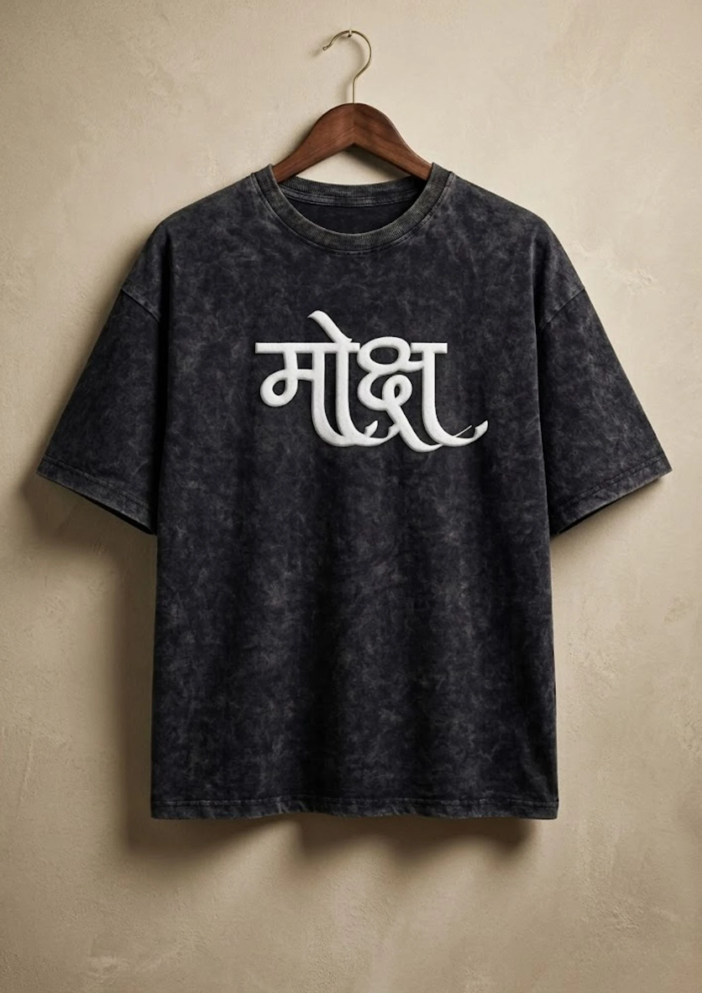 Oversized black acid wash t-shirt with 3D puff print Hindi "Moksha" (मोक्ष) calligraphy graphic in white, hanging on a wooden hanger against a beige wall background