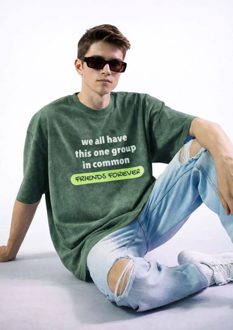 Acid wash olive green oversized t-shirt with white and neon green typography reading "We All Have This One Group In Common — Friends Forever" floating against a grey background